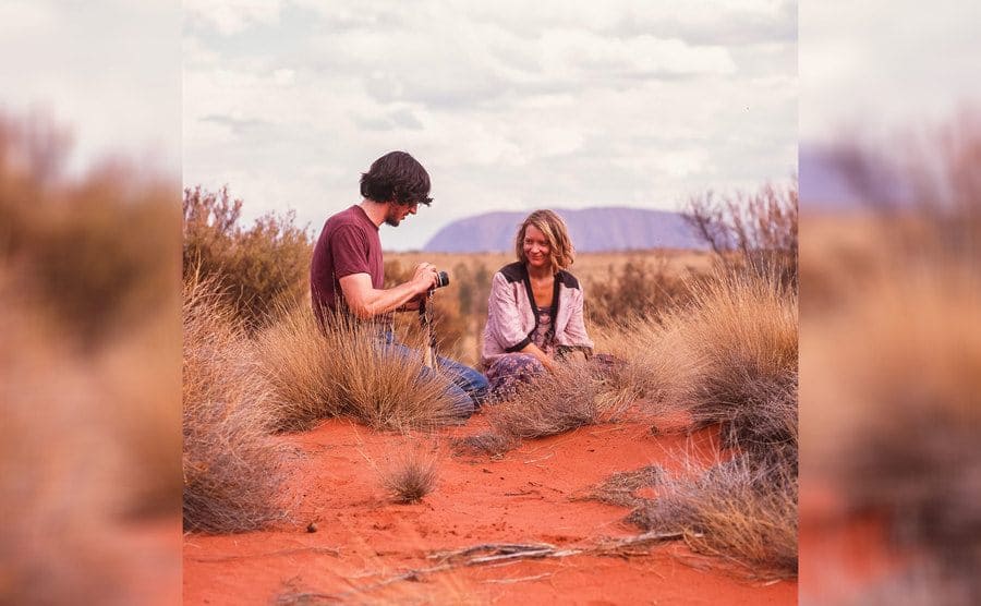 Mia Wasikowska and Adam Driver are sitting in the desert while Adam toys with his camera in the film Tracks. 