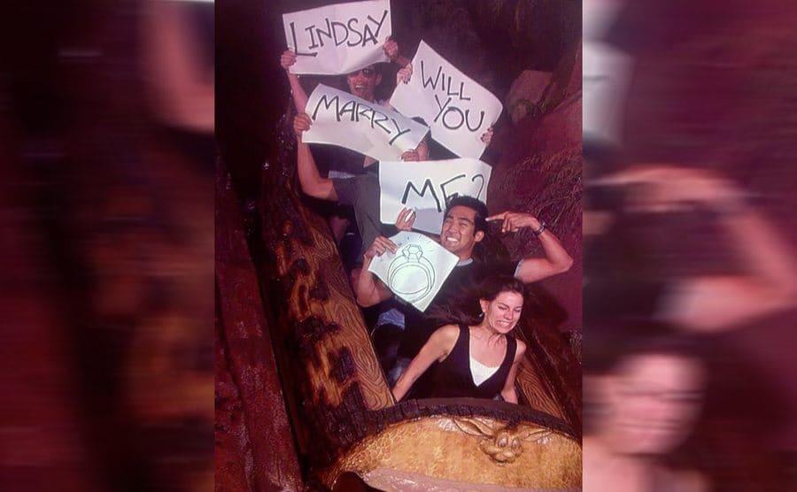Four men holding up signs that say, “Lindsay will you marry me?” with a drawing of an engagement ring and a woman sitting in the front looking very scared 