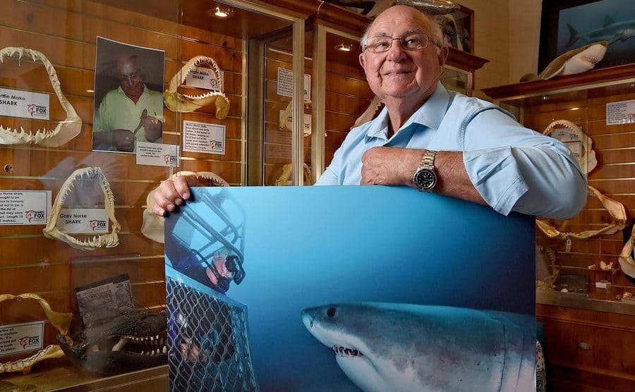 Rodney Fox posing in front of Shark jaws mounted in cases while he holds a photo of himself in a cage looking out at a shark 